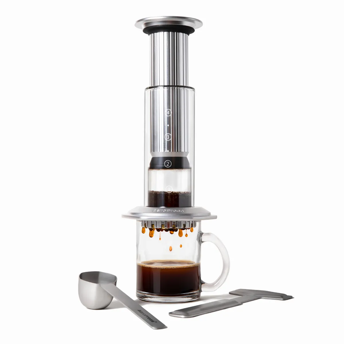 AeroPress Premium in silver pressing coffee into a glass mug, with its steel scoop and stirrer
