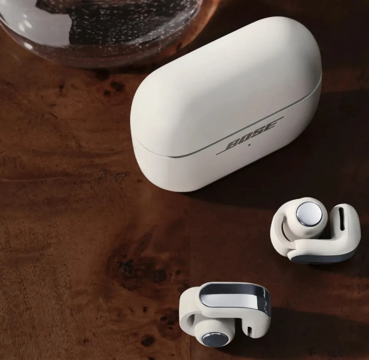 The Bose Ultra Open Earbuds (2nd Gen) in White Smoke next to their charging case on a wooden table